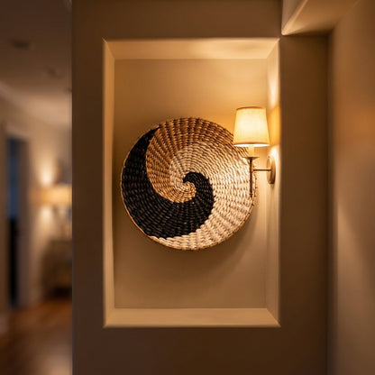 Round woven wall basket with a black, tan and cream swirl pattern hung in a recessed wall niche beside a lit sconce with a cream drum shade