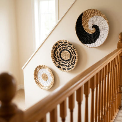 Three round woven wall baskets in black, white and tan patterns stepping up a cream wall beside an oak staircase banister