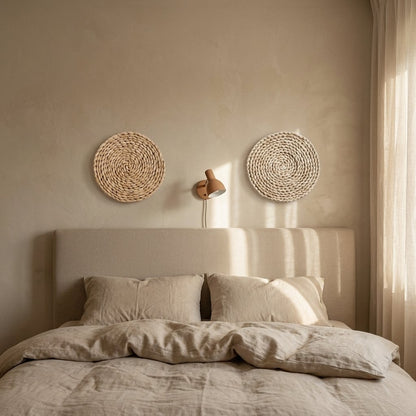 Two flat natural tan coiled woven wall discs hung above a bed with an oatmeal headboard, with a small terracotta swing-arm lamp between them