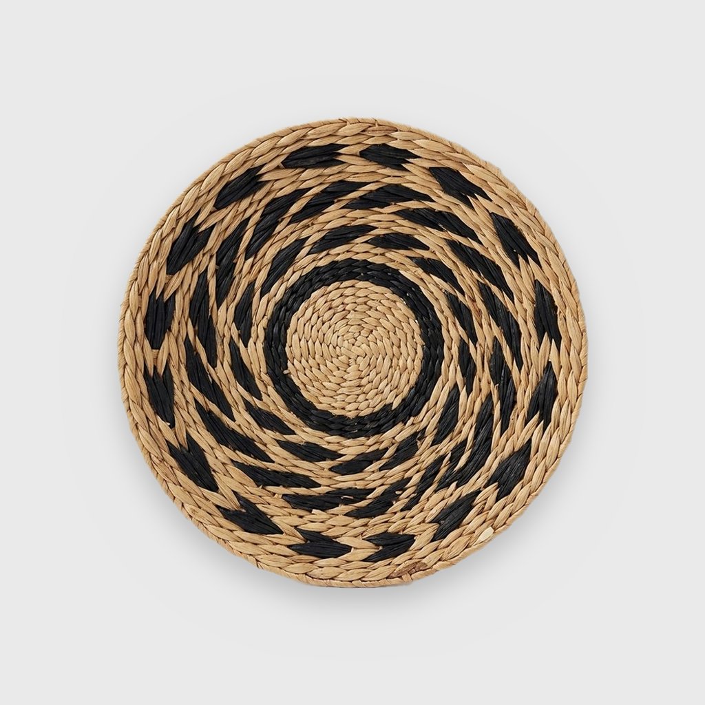 Single round woven basket with a tan center, a black ring and black leaf-shaped dashes on tan in a swirling pattern, on a plain light gray background