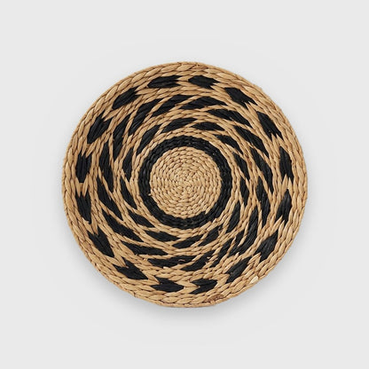 Single round woven basket with a tan center, a black ring and black leaf-shaped dashes on tan in a swirling pattern, on a plain light gray background
