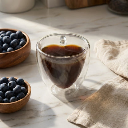 Heart-shaped glass mug filled with coffee on a marble countertop beside bowls of fresh blueberries and a folded linen napkin