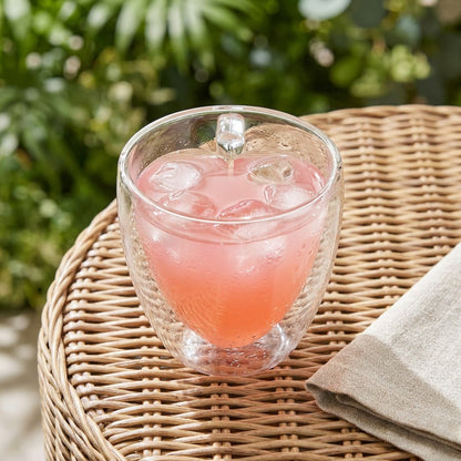 Heart-shaped glass mug filled with a pink iced drink and ice on a wicker table outdoors