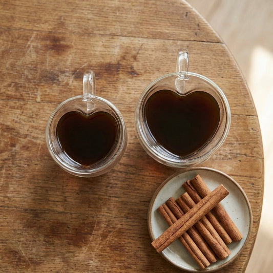 Small and large heart-shaped glass mugs filled with coffee, shown side by side from overhead with a small plate of cinnamon sticks