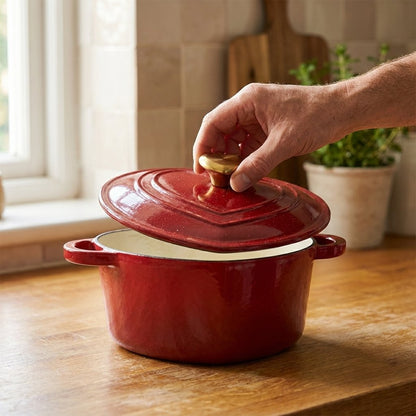 Hand lifting the lid off a red heart-shaped enamel pot by its gold heart knob, revealing the cream enamel interior