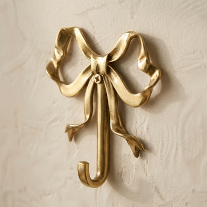 Close-up of a gold bow-shaped wall hook with a screw at the knot, mounted on a cream plaster wall