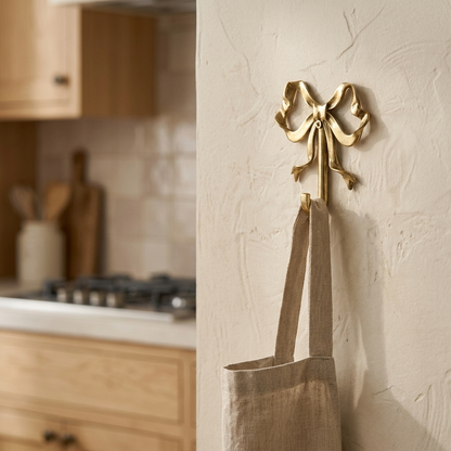 Gold bow-shaped wall hook holding a linen tote on a plaster wall beside a kitchen cooktop