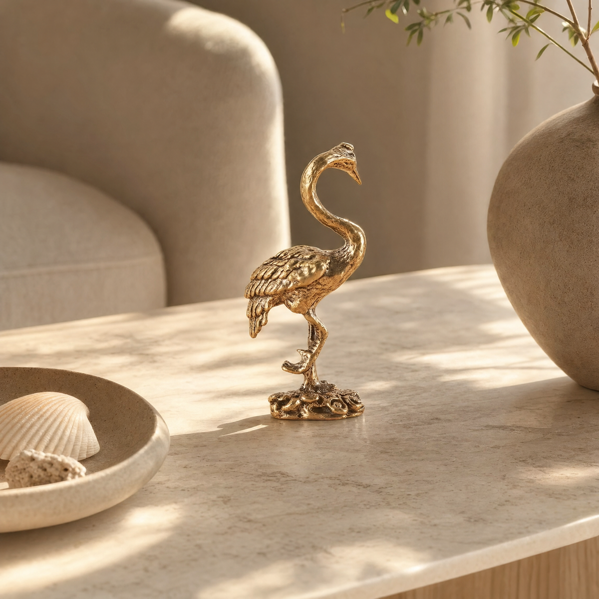 Gold brass crane figurine standing on one leg on a pale marble tabletop beside a large round gray-beige vase and a cream dish holding a scallop shell and coral