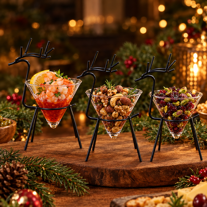 Three black reindeer stand cone glasses filled with a shrimp cocktail and two nut and dried fruit snack mixes on a wooden serving board with pine branches and Christmas lights
