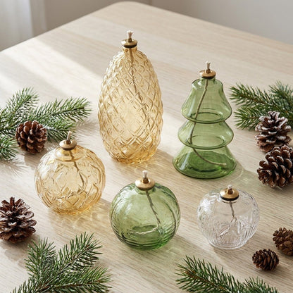 Five styles of Holiday Glass Oil Lamps, Pinecone, Gold Ornament, Tree, Green Ornament, and Clear Ornament, displayed together on a table with pine branches