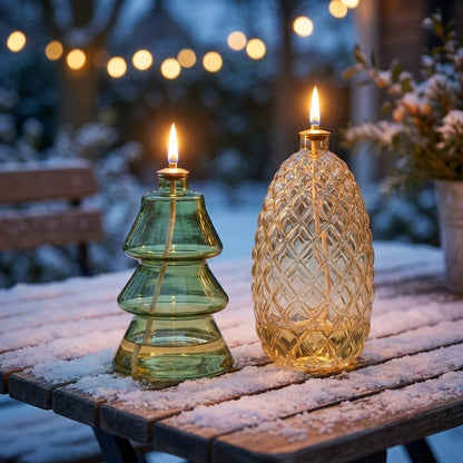 Lit Tree and Pinecone glass oil lamps on a snow-dusted patio table at dusk with string lights