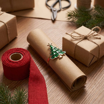 Green enamel Christmas tree napkin ring with gold trim resting on kraft gift wrapping paper beside ribbon and twine