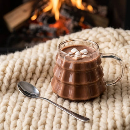 Tree-shaped glass filled with hot cocoa and marshmallows on a knit blanket beside a lit fireplace