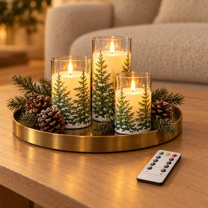 Three flameless candles printed with snowy pine trees on a round brass tray with pinecones and evergreen sprigs on a wood coffee table, with a remote control beside it and a cream sofa behind
