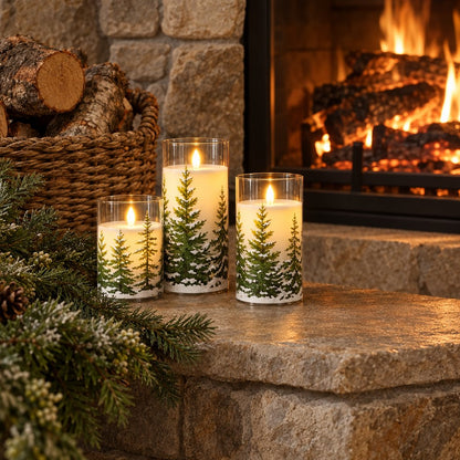Three flameless candles printed with snowy pine trees on a stone hearth with frosted evergreen sprigs, beside a lit fireplace and a basket of logs