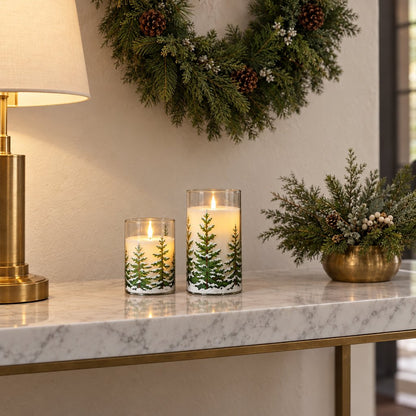Two flameless candles printed with snowy pine trees on a marble console beside a table lamp, a small gold pot of evergreen sprigs and a pine wreath on the wall