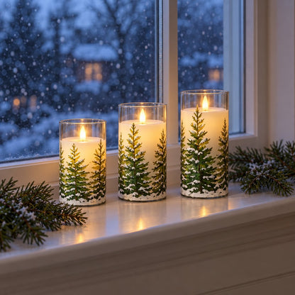 Three flameless candles printed with snowy pine trees on a window sill with evergreen sprigs, in front of a window with snow falling outside at dusk