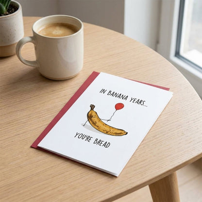Greeting card with banana illustration on a wooden table with a mug of coffee.