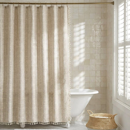 Natural oatmeal windowpane linen shower curtain with small cream tassels beside a clawfoot tub and woven basket in a bright cream-tiled bathroom