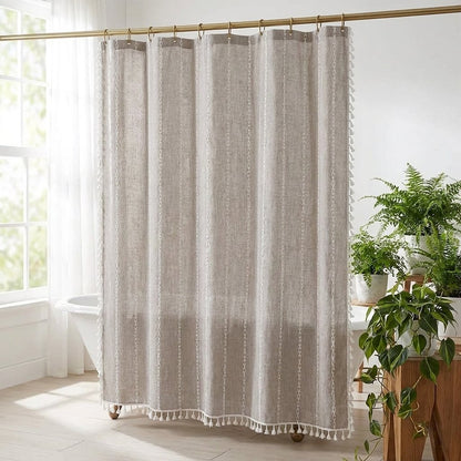Oatmeal linen shower curtain with thin cream vertical stripes and white tassels on a brass rod in a bright bathroom with a clawfoot tub and ferns