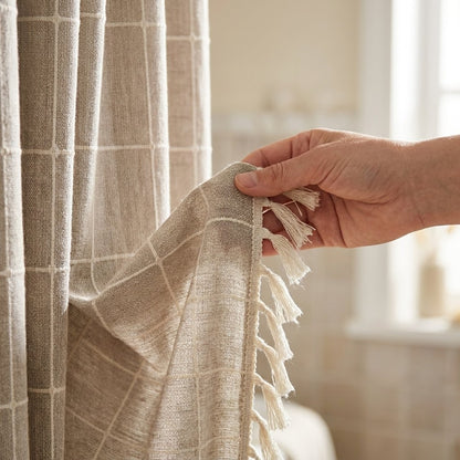 Hand holding the edge of an oatmeal windowpane linen shower curtain to show the cream tassels along the side