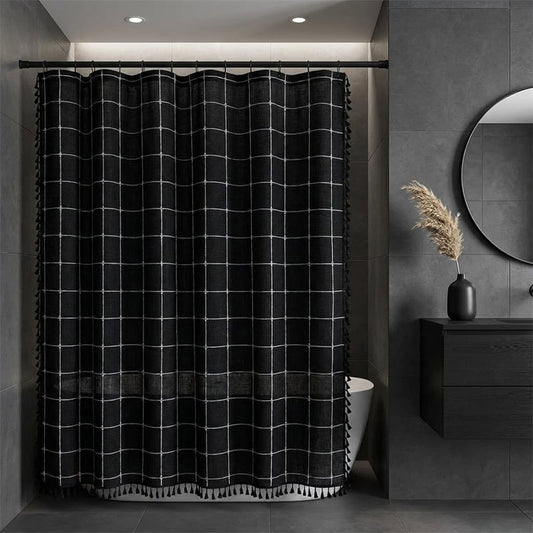 Black linen shower curtain with a thin white windowpane grid and tassels along the sides and hem in a dark gray-tiled bathroom with a round mirror and pampas grass