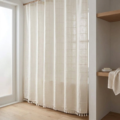 Cream semi-sheer windowpane linen shower curtain with a small tassel hem in a minimalist bathroom with light oak flooring and a shelf with a towel and soap dish