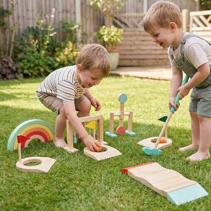Two young boys playing with the wooden mini golf set in a backyard, putting a ball toward a flagged hole beside the rainbow arch piece