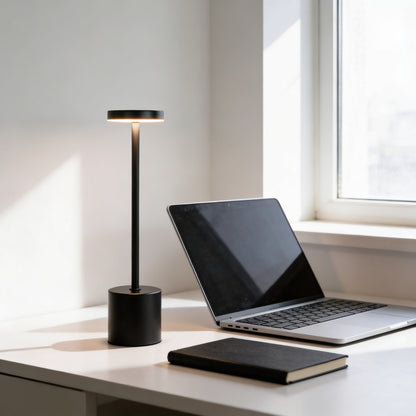 Laptop on a desk with a black lamp and notebook in a bright room.