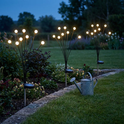 Three lit solar garden lights along a stone-edged flower border beside a galvanized watering can at dusk