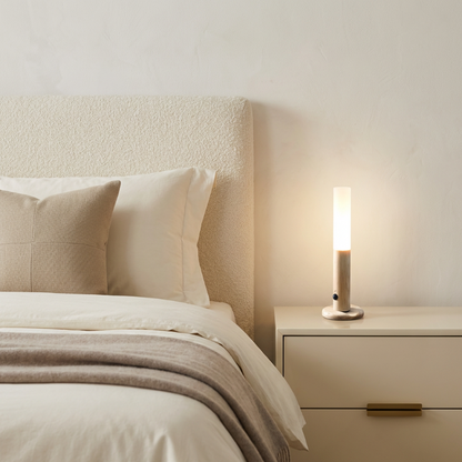 Neatly made bed with beige pillows and blanket, nightstand with lamp in a softly lit bedroom.