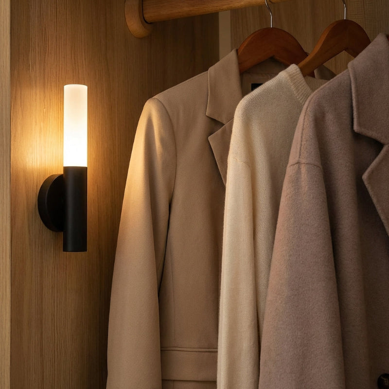 Three coats hanging on a rack with a wall sconce casting light on them.