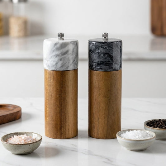 White and black marble-topped acacia wood salt and pepper mills beside small bowls of salt and peppercorns on a marble counter