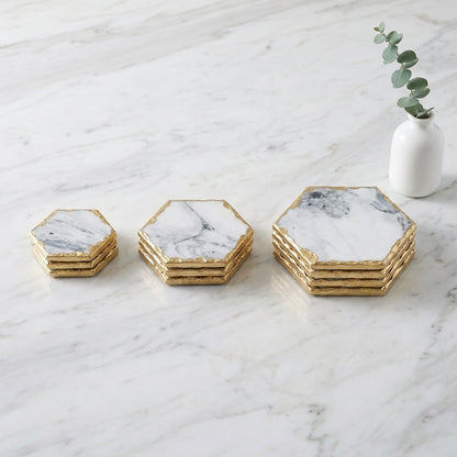 Stacks of small, medium and large hexagonal white marble coasters with gold edging on a white marble countertop beside a vase with eucalyptus