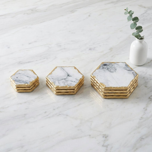 Stacks of small, medium and large hexagonal white marble coasters with gold edging on a white marble countertop beside a vase with eucalyptus