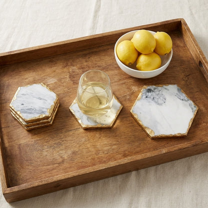 Small, medium and large hexagonal marble coasters with gold edging on a wooden tray with a glass of white wine and a bowl of lemons