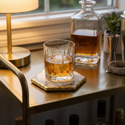 Hexagonal white marble coaster with gold edging under a glass of whiskey with ice on a brass bar cart beside a decanter