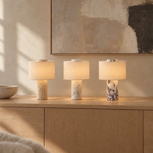 Three lit portable table lamps with cream linen drum shades on an oak credenza, with travertine, white marble and burgundy-veined marble bases