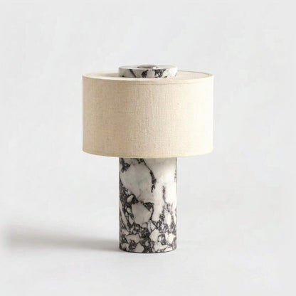 Portable table lamp with a white marble base and bold dark grey veining and a beige linen drum shade on a plain light backdrop