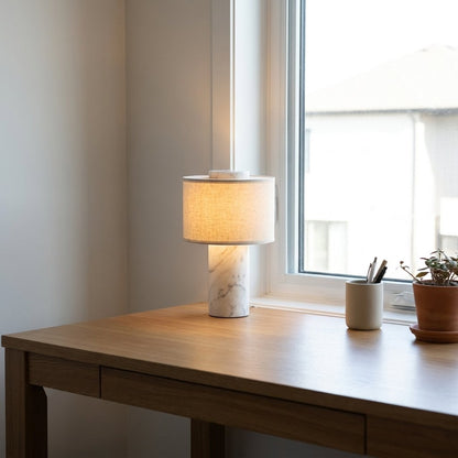 Lit portable table lamp with a white marble base on an oak desk by a window beside a cream pen cup and a small potted plant