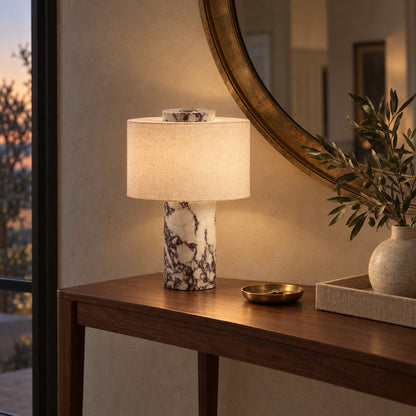 Lit portable table lamp with a burgundy-veined white marble base on a dark walnut console with a round brass mirror and an olive branch in a vase
