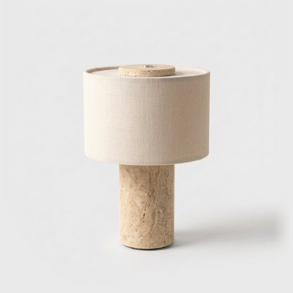 Portable table lamp with a warm beige pitted travertine base and a cream drum shade on a plain light backdrop