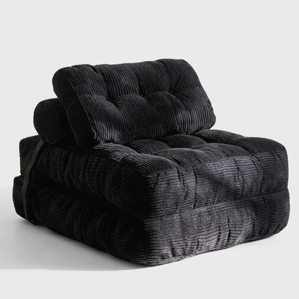 A black corduroy tufted floor cushion or small chair with a matching rolled cushion on top, photographed on a clean light gray studio background.