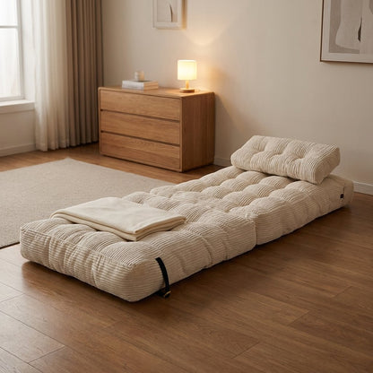 A cream-colored ribbed floor mattress topper with an integrated pillow, styled on a wooden floor next to a matching folded blanket in a warm, modern bedroom setting.