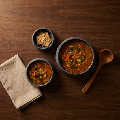 Overhead view of three dark blue-gray stoneware bowls, two with lentil and vegetable soup and one with crackers, on a dark wood table