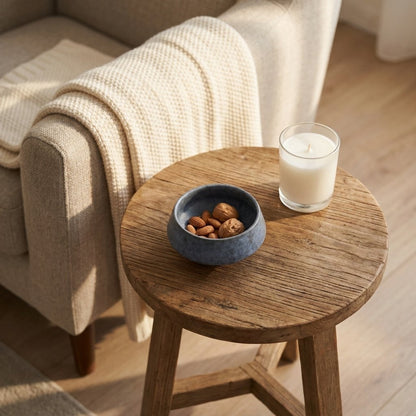 Small blue stoneware bowl of mixed nuts on a round wooden side table beside a candle and an armchair with a knit throw