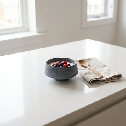 Small matte blue stoneware bowl of yogurt topped with raspberries and blueberries on a white counter beside a wooden spoon