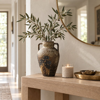 Tan and cream two-handled ceramic vase with a dark brown patch holding olive branches on an oak console beside a lit candle, a brass tray and a round gold mirror