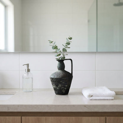 Dark charcoal-green ceramic pitcher vase with a single handle holding eucalyptus stems on a stone bathroom counter beside a glass pump bottle and a folded white towel