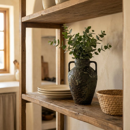 Dark green-charcoal two-handled ceramic vase holding eucalyptus stems on a weathered wood shelf beside a stack of cream plates and a woven basket